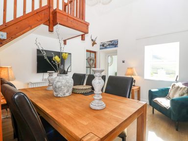 A dining area with a wooden table black chairs a teal armchair and a wall-mounted TV at Pennant Cottage in Llithfaen
