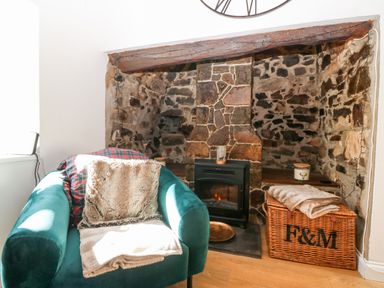 A green armchair with blankets in front of a stone fireplace with a stove and a wicker basket at Pennant Cottage in Llithfaen