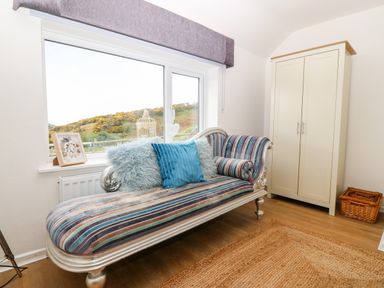 A room with a striped chaise lounge and cushions near a window a cream wardrobe and a wicker basket on a wooden floor at Pennant Cottage in Llithfaen