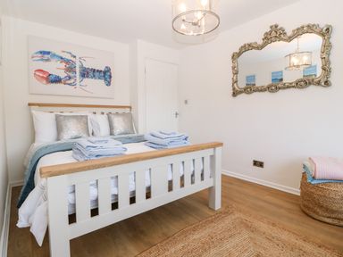 A bedroom with a white wooden bed folded towels and blankets on a wicker basket a decorative mirror on the wall and a lobster artwork at Pennant Cottage in Llithfaen