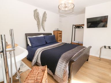 A bedroom with a bed a freestanding bathtub a wooden chest of drawers a clothing rack and a wall-mounted TV at Pennant Cottage in Llithfaen