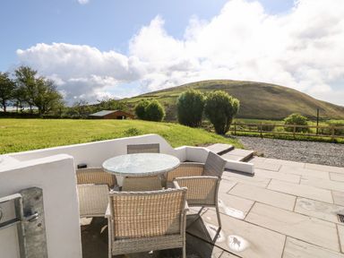 An outdoor patio with a round glass table and four wicker chairs overlooking a grassy hill and trees at Pennant Cottage in Llithfaen
