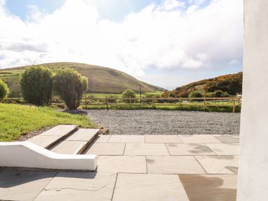 A paved patio with steps leading to a gravel area with bushes and a wooden fence with hills in the background at Pennant Cottage in Llithfaen