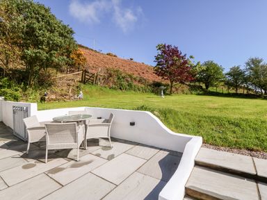 An outdoor patio with a glass table and four chairs overlooking a grassy garden with trees and a bird bath at Pennant Cottage in Llithfaen