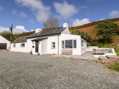 A white cottage with a gravel driveway patio with outdoor furniture and a hillside in the background at Pennant Cottage in Llithfaen