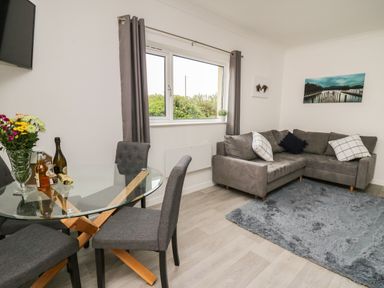 A living room with a glass dining table and grey chairs next to a grey corner sofa and a blue rug at Ty Cariad in Llangynin near St Clears