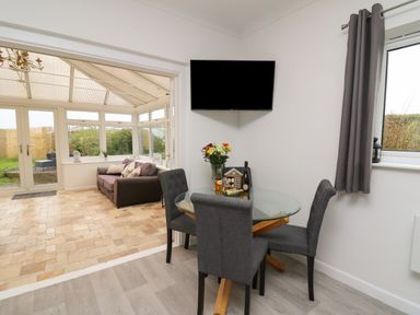 A dining area with a glass table and four chairs with a TV mounted on the wall and a conservatory with a sofa and glass door at Ty Cariad in Llangynin near St Clears
