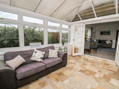 A conservatory with a purple sofa and tiled floor opening to a dining area with gray chairs and a living room at Ty Cariad in Llangynin near St Clears