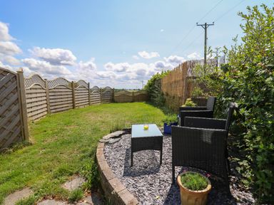 A garden area with two wicker chairs and a glass table on a stone surface next to a wooden fence and plants at Ty Cariad in Llangynin near St Clears