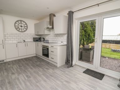 A kitchen with white cabinets a wall clock and glass doors leading to a garden with outdoor furniture at Ty Cwtch in Llangynin near St Clears