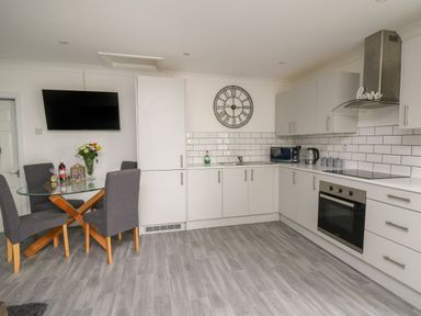 A kitchen with white cabinets, a wall clock, a glass dining table with four chairs, and a wall-mounted TV at Ty Cwtch in Llangynin near St Clears