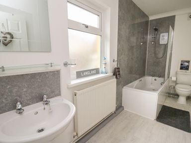 A bathroom with a sink, bathtub with shower, toilet, radiator, window with a Langland sign and a lighthouse decoration at Ty Cwtch in Llangynin near St Clears