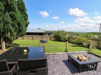 An outdoor patio with a glass table and chairs a fire pit and a grassy fenced garden with trees at Ty Cwtch in Llangynin near St Clears