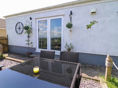 An outdoor patio with a glass table and wicker chairs by white double doors with hanging plants and wall decorations at Ty Cwtch in Llangynin near St Clears