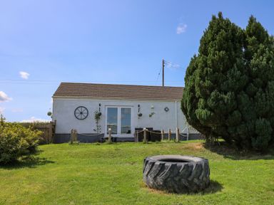 A small white building with double doors and outdoor seating on a grassy area near large evergreen trees and a large tire in the foreground at Ty Cwtch in Llangynin near St Clears
