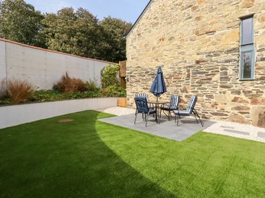 An outdoor patio area with a table and four chairs with cushions and an umbrella on a stone-paved section next to a stone building and green lawn at Carines Barns - Pol in Cubert