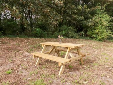 A wooden picnic table with a small vase of flowers on top outdoors near trees at Carines Barns - Pol in Cubert