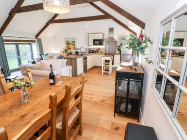 An open plan kitchen and dining area with wooden dining table and chairs a kitchen island white cabinets sofa and large window at Carines Barns - Pol Cubert