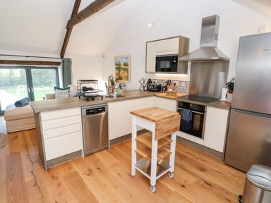 A kitchen with wooden floors a small movable island stainless steel appliances and a living area with windows at Carines Barns - Pol in Cubert
