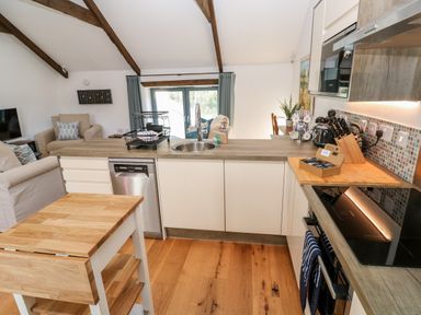 A kitchen with wooden floors, white cabinets, a sink, stove, and a wooden island with a living area visible in the background at Carines Barns - Pol in Cubert