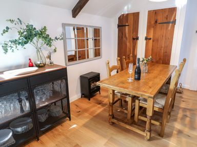 A dining room with a wooden table and chairs a bottle and glass on the table a black cabinet with dishes and a vase with greenery on top a wall mirror and wooden cupboard doors at Carines Barns - Pol in Cubert