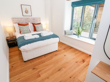 A bedroom with a bed with pillows and folded towels bedside tables with lamps and a window seat with cushions and a plant at Carines Barns - Pol in Cubert