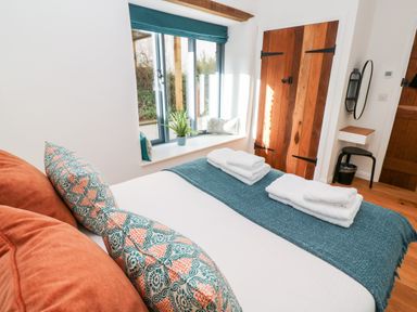 A bedroom with a bed with folded towels pillows and a blue throw near a window with cushions and wooden doors at Carines Barns - Pol in Cubert