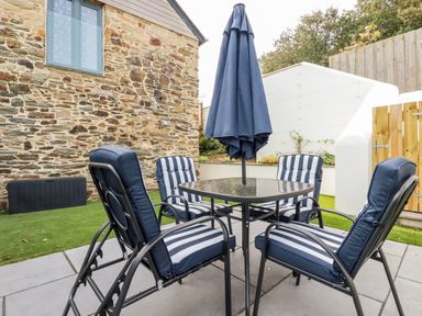An outdoor patio area with a stone wall two striped cushioned chairs two cushioned recliners a glass table with a closed umbrella at Carines Barns - Par in Cubert