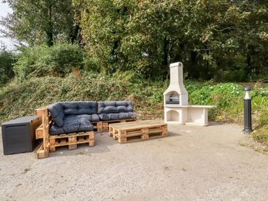 An outdoor seating area with wooden pallet furniture and a barbecue grill surrounded by greenery at Carines Barns - Par in Cubert