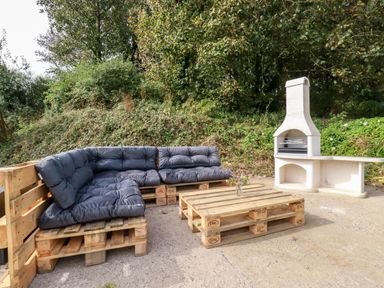 An outdoor seating area with wooden pallet furniture and a white stone barbecue surrounded by greenery at Carines Barns - Par in Cubert