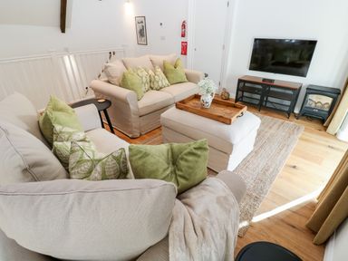 A living room with two sofas, green cushions, an ottoman with a wooden tray, a wall mounted TV and a small stove at Carines Barns - Par in Cubert