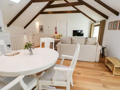 A dining area with a round white table and chairs and a living area with beige sofas and a wall-mounted TV in a room with wooden ceiling beams at Carines Barns - Par in Cubert