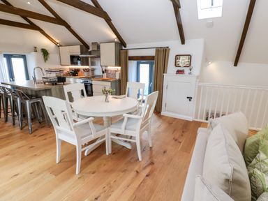 An open kitchen and dining area with wooden beams white dining table and chairs and a sofa at Carines Barns - Par Cubert