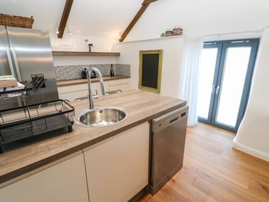A kitchen with a wooden countertop island with a round sink and chrome faucet a stainless steel dishwasher cream cabinets a stainless steel refrigerator open shelves with glassware a tiled backsplash and double glass doors at Carines Barns - Par in Cubert
