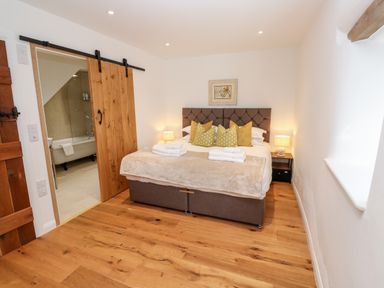 A bedroom with a double bed pillows bedside tables lamps wooden floor and a sliding wooden door leading to a bathroom at Carines Barns - Par in Cubert