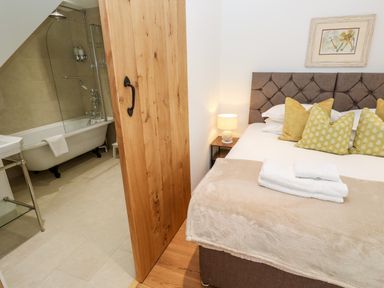 A bedroom with a bed and bedside table and a bathroom with a bathtub seen through a wooden sliding door at Carines Barns - Par in Cubert