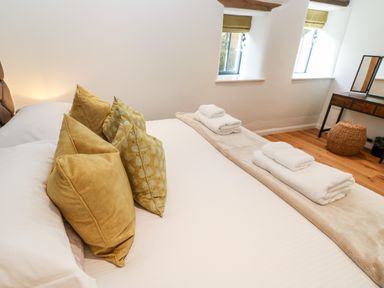 A bedroom with a bed with yellow pillows and folded towels on a beige runner by windows and a wooden vanity at Carines Barns - Par in Cubert