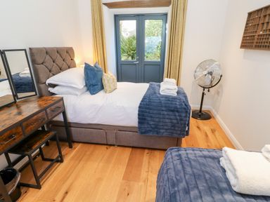 A bedroom with two single beds a wooden desk a standing fan and double doors with yellow curtains at Carines Barns - Par in Cubert