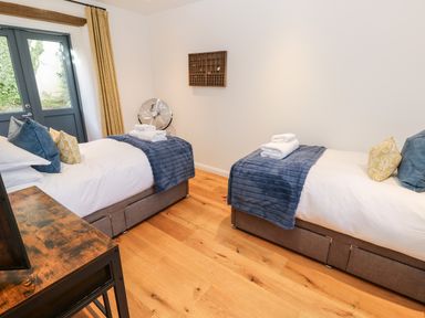 A bedroom with two single beds with blue throws and folded towels next to a window with mustard curtains at Carines Barns - Par in Cubert