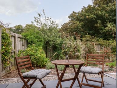 A small outdoor patio with two wooden chairs with cushions and a round wooden table with a glass vase surrounded by plants and a wooden fence at Carines Barns - Wesley Barn Cubert
