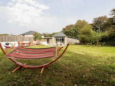 A garden with a striped hammock wooden outdoor dining set and stone building in the background at Carines Barns - Wesley Barn Cubert