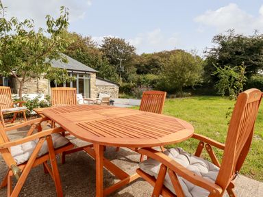 An outdoor patio with a wooden table and chairs next to a stone building and green lawn at Carines Barns - Wesley Barn Cubert