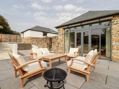 An outdoor patio with wooden cushioned chairs a table and a fire pit by a stone building with large windows at Carines Barns - Wesley Barn in Cubert