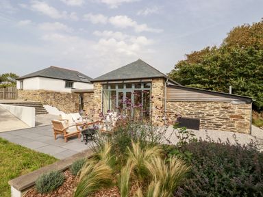 A stone building with large windows patio area with wooden chairs and table garden plants at Carines Barns - Wesley Barn in Cubert
