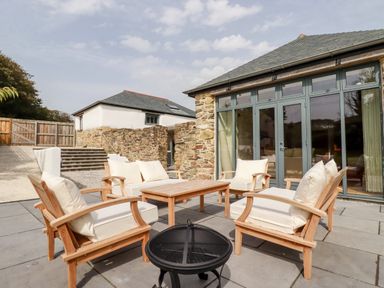 An outdoor patio with wooden chairs and a table next to a stone building with large glass doors at Carines Barns - Wesley Barn in Cubert