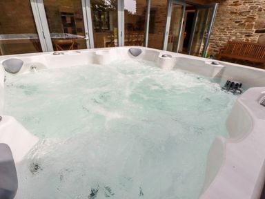 A bubbling hot tub in an outdoor area with stone walls glass doors and a wooden bench at Carines Barns - Wesley Barn in Cubert