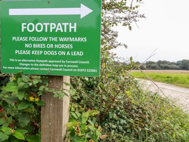 A green footpath sign on a wooden post next to a hedgerow with a dirt path and fields in the background at Carines Barns - Wesley Barn Cubert