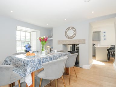 A dining area with a table covered by a patterned tablecloth set with plates and glasses near a window and a kitchen at Cob Cottage near Rock