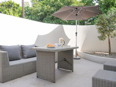 An outdoor patio with a grey wicker sofa table umbrella and two stools at Cob Cottage near Rock