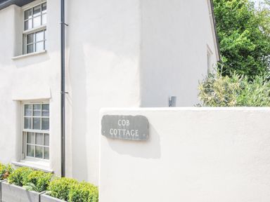 The exterior corner of a white cottage with windows plants and a sign reading Cob Cottage near Rock